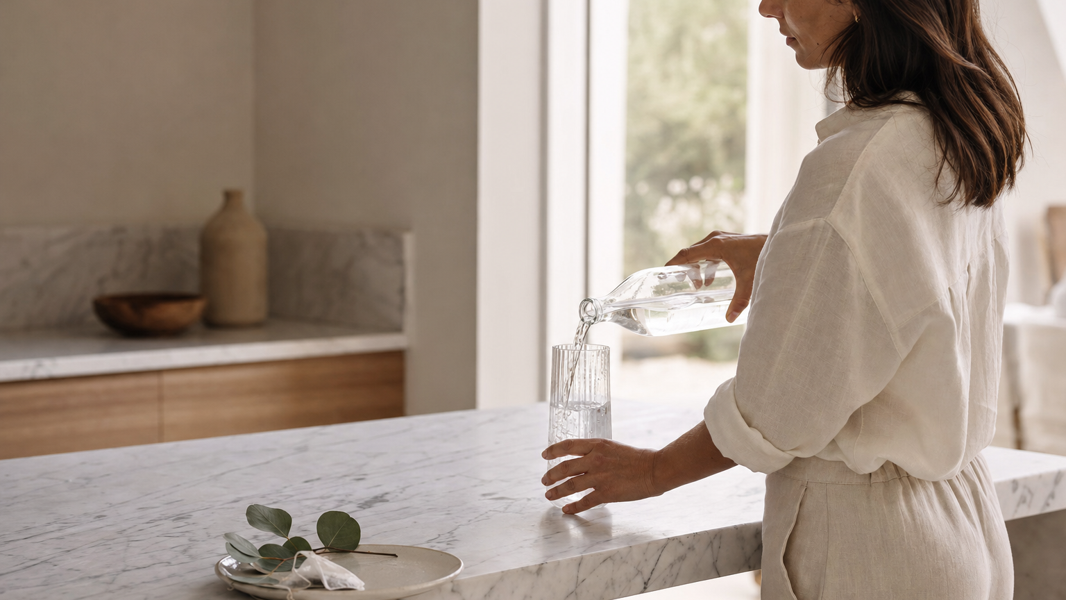 Mujer vertiendo agua mineral en vaso de cristal sobre mármol — rutina mañana premium